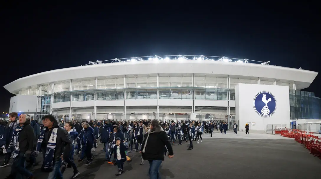 Stadion Tottenham Hotspur nocą, biały fasada z herbem koguta, kibice w granatowo-białych szalika przechodzą obok