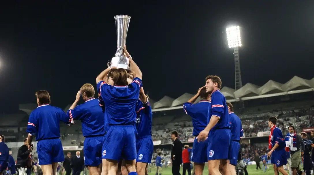 Les joueurs du Paris Saint-Germain, en maillots bleu royal, tiennent en l'air un grand trophée en argent sous un pylône de projecteurs la nuit