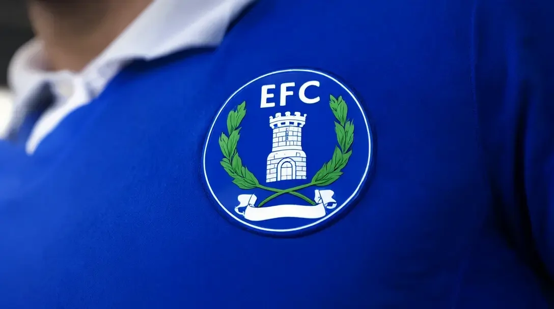Close-up do escudo do Everton em uma camisa azul real: torre branca, grinal verde, letras EFC e um pergaminho em branco