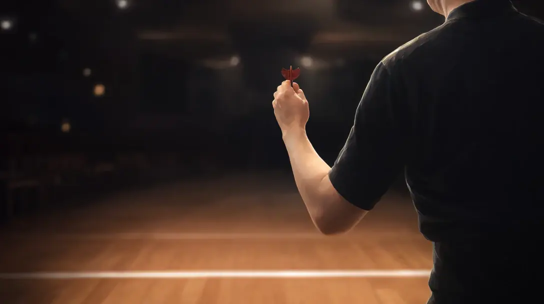 Anonymous darts player seen from behind mid-throw on a stage, in a dark hall with warm spotlights.