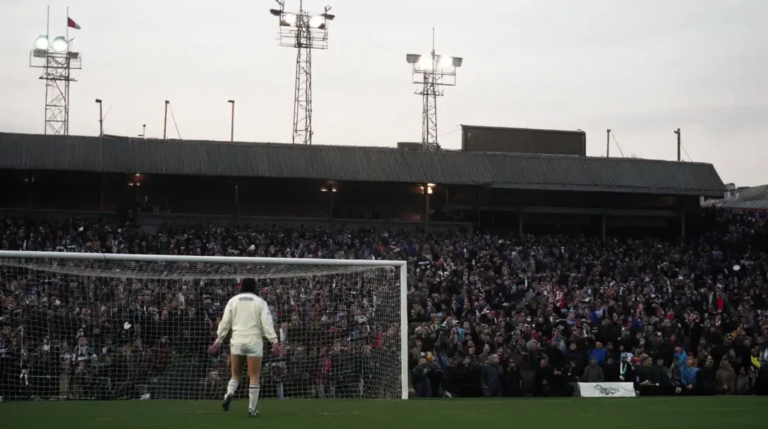Goleiro com camisa creme e luvas rosa visto de costas na meta, arquibancada lotada e torres de iluminação ao fundo