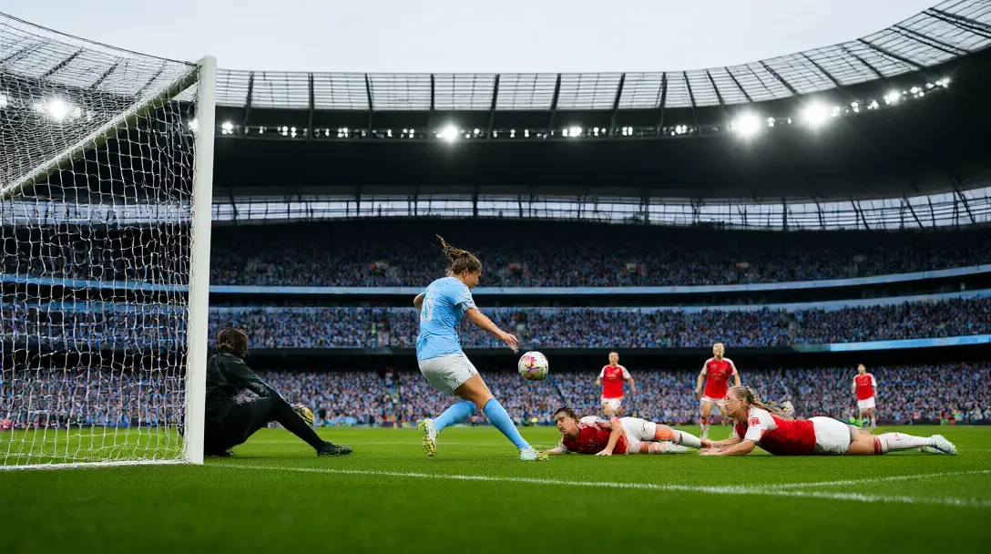 Manchester City forward in sky blue shooting past a diving goalkeeper at the Etihad as Arsenal defenders fall away