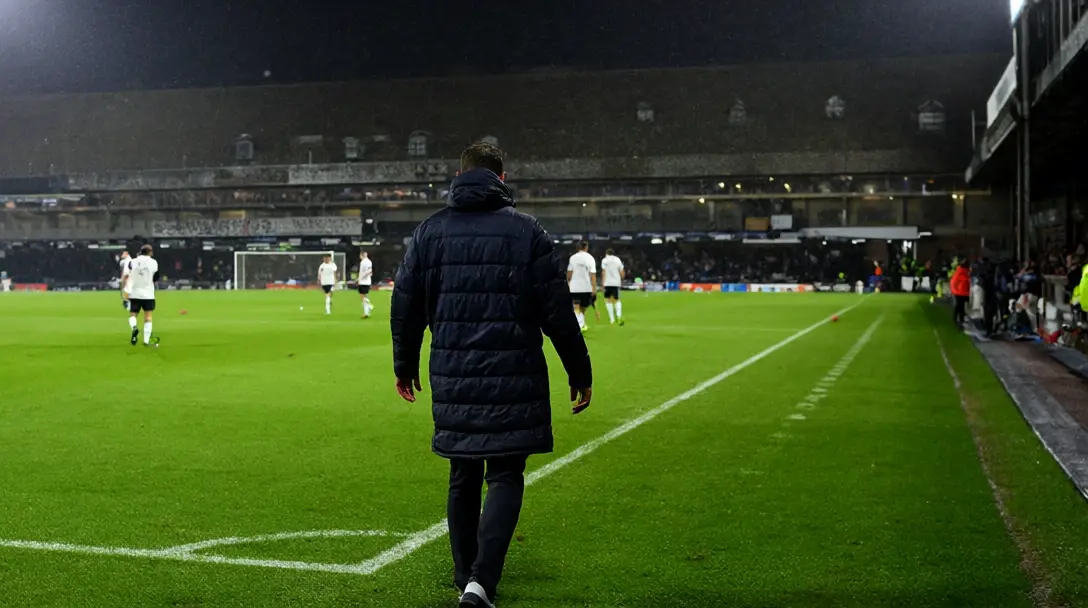 Manager in a dark padded coat seen from behind on the touchline of a floodlit pitch, players in white shirts warming up beyond.