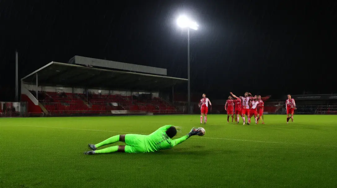 Lower-league goalkeeper diving to save under floodlights at a small English ground on a wet night
