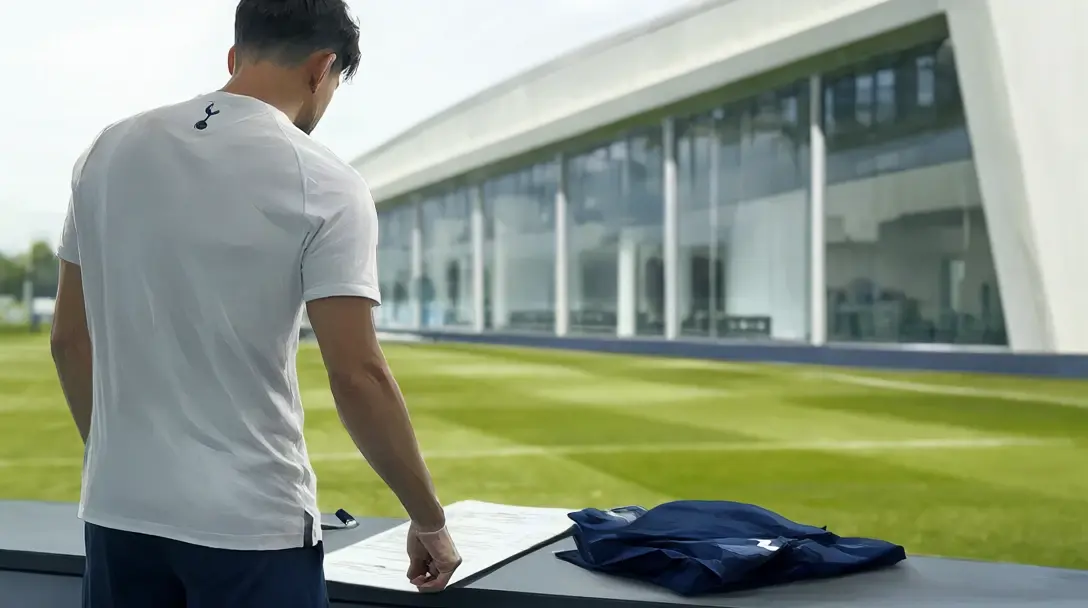 Footballer in white shirt and navy shorts seen from behind leaning over a contract at a training centre, navy top on the table