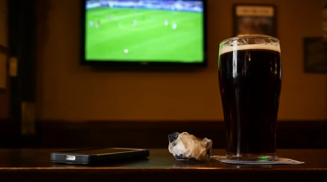 Pub table with a pint and a face-down phone, a football match on the TV blurred in the background