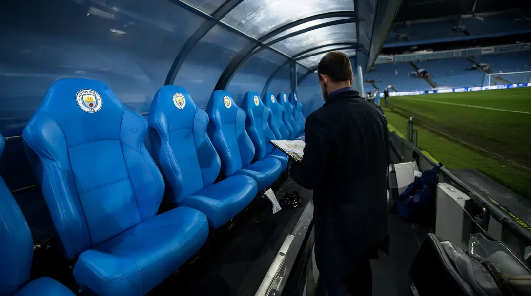 Manchester City dugout with sky-blue seats bearing the club crest, a man in a dark coat reading papers beside the pitch