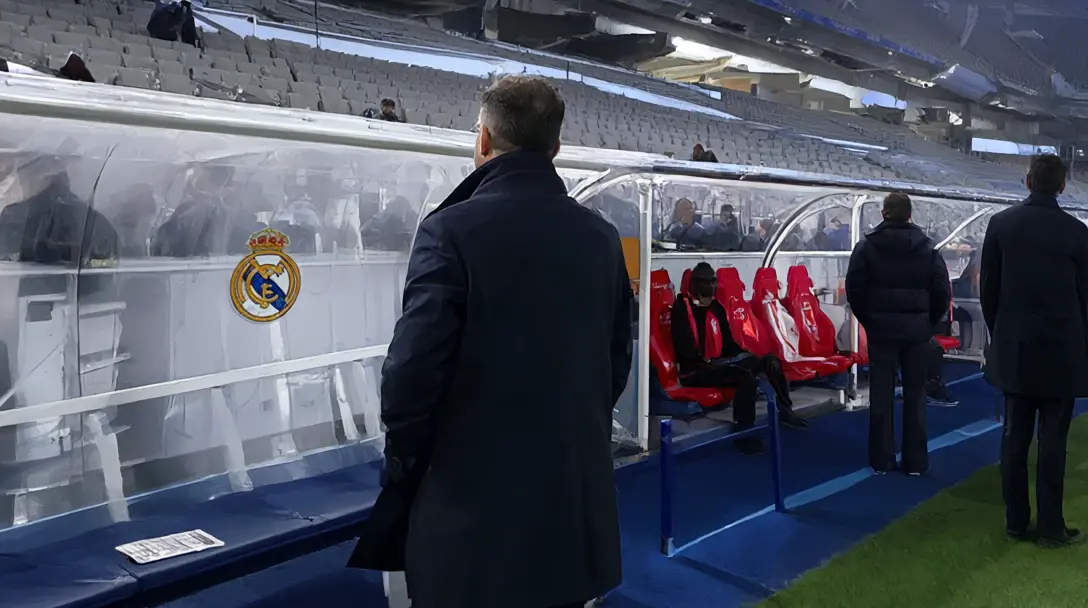 Manager in a dark overcoat seen from behind beside the Real Madrid dugout with its crest, in an empty stadium.