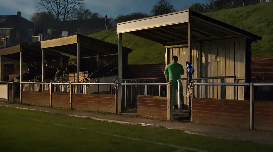 Small English football ground at dusk with a goalkeeper in plain green kit seen from behind.
